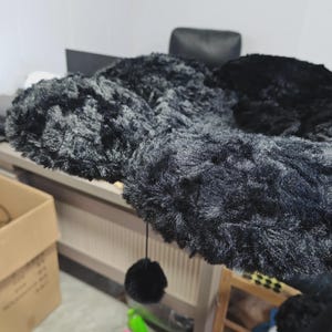 May include: A black cat tree with multiple platforms covered in soft, plush material. A dangling black pom-pom toy hangs from one of the platforms. The cat tree is designed for indoor use and provides a space for cats to play and rest.
