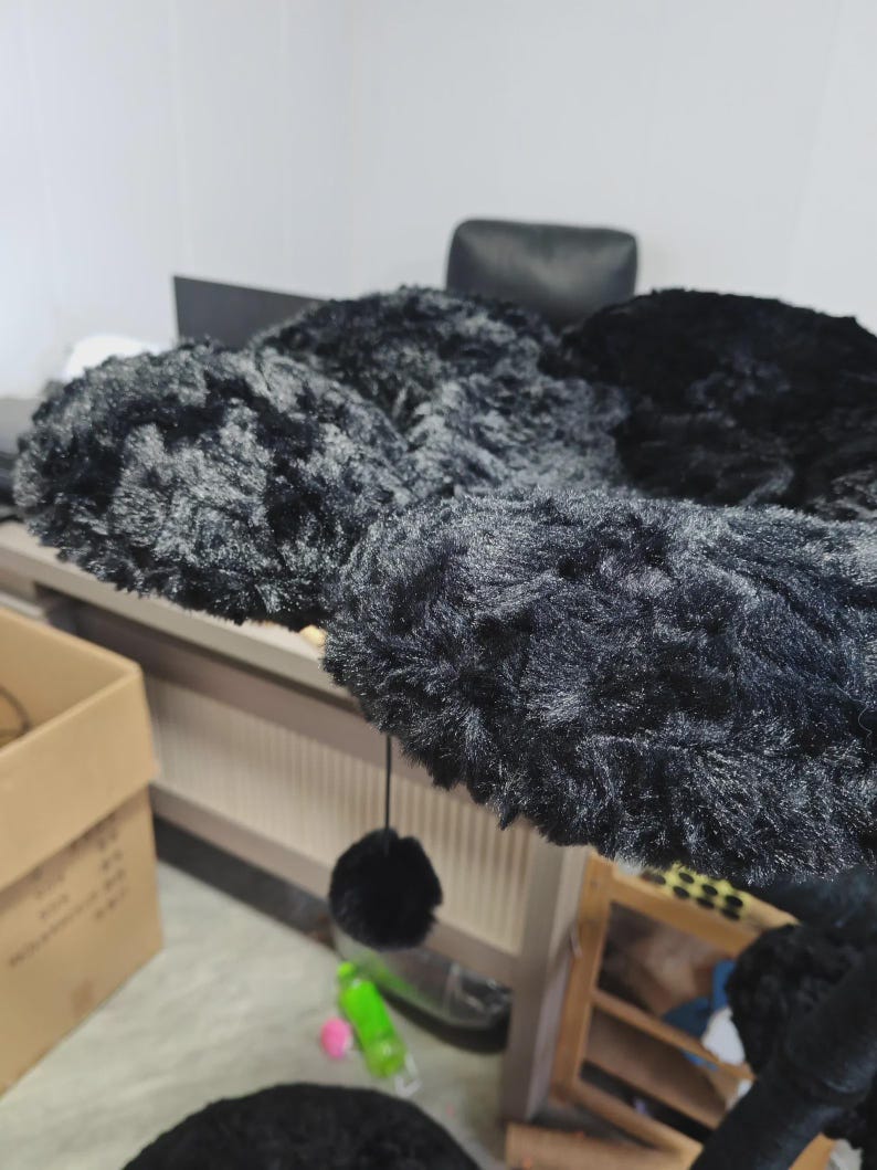 May include: A black cat tree with multiple platforms covered in soft, plush material. A dangling black pom-pom toy hangs from one of the platforms. The cat tree is designed for indoor use and provides a space for cats to play and rest.