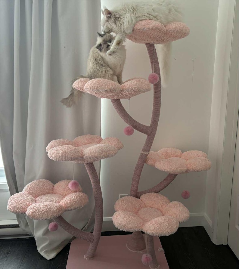 May include: A pink cat tree with flower-shaped platforms and dangling pom-poms. Two fluffy cats are on the tree. The tree is made of a soft, plush material, providing a comfortable and engaging space for cats. The tree is in a room.