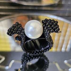 May include: A black beaded ring with a large, round, white pearl center. The ring has a unique design with beaded accents around the pearl. The ring is displayed on a reflective surface.