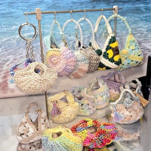 May include: A collection of handmade crochet bags in various colors and designs. The bags are displayed on a gold rack and a reflective surface. Some bags have handles, tassels, and keychains. The color palette includes pastels, brights, and neutral tones.
