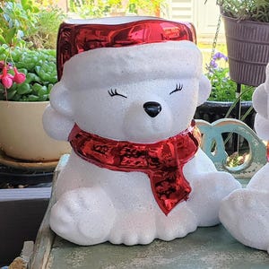 Op de afbeelding: Een witte ijsbeerfiguur met een rode kerstmuts en sjaal. De beer heeft gesloten ogen en een zwarte neus. Het beeldje is bedekt met glitter en staat op een lichtblauwe ondergrond. De achtergrond bevat groen.