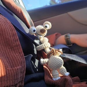 Op de afbeelding: Gehaakte Sid de Luiaard en een kleiner gehaakt dier, mogelijk een hond, zijn bevestigd aan een veiligheidsgordel. Sid de Luiaard heeft grote, wiebelende ogen en een bruine snuit. De achtergrond toont een auto-interieur en een glimp van de lucht.