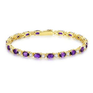 Pode incluir: Pulseira dourada com pedras preciosas roxas de corte oval e pequenas pedras brancas redondas. A pulseira tem um fecho seguro e um padrão repetitivo de pedras preciosas. A joia está sobre um fundo branco.