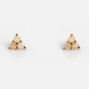 Könnte beinhalten: Ein Paar goldene Ohrstecker, jeweils mit einer dreieckigen Anordnung aus drei opalisierenden Edelsteinen. Die Ohrringe sind vor einem weißen Hintergrund platziert, was ihr zartes Design und die subtilen Farbvariationen hervorhebt.