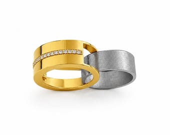 May include: A two-piece interlocking ring set featuring a polished gold band with a horizontal row of small clear stones and a second band with a textured, matte grey finish. The gold band has a wide, flat profile with a cutout section.