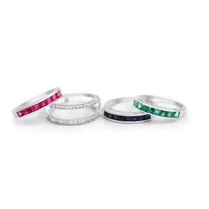 May include: A collection of silver rings with colorful gemstones. One ring features a row of red gemstones, another has clear stones, and others have blue and green stones. The rings are displayed on a white background.