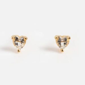 Puede incluir: Un par de pendientes de botón dorados, cada uno con una piedra preciosa clara en forma de corazón. Los pendientes tienen un diseño sencillo y elegante, perfecto para el uso diario. Las piedras preciosas están facetadas, captando la luz y añadiendo un toque de brillo.