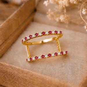 May include: A gold double-band ring featuring two parallel bars connected by vertical gold struts. Each bar is set with a continuous row of alternating round-cut pink rubies and clear white diamonds, creating a geometric and symmetrical design.