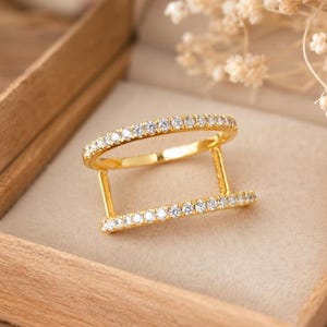 Peut inclure: Une bague dorée composée de deux anneaux parallèles reliés par deux barres verticales. Les deux bandes horizontales sont incrustées d'une rangée de petites pierres claires taillées en rond qui captent la lumière, offrant un aspect structuré.