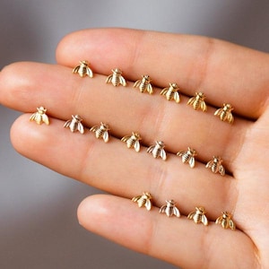 Op de afbeelding: Een verzameling kleine, goudkleurige oorstekers in de vorm van bijen. De oorbellen zijn in rijen op een hand gerangschikt en tonen hun delicate ontwerp en metallic glans. De bijen-oorstekers zijn een charmant accessoire.