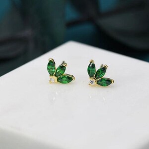 Op de afbeelding: Een paar goudkleurige oorstekers, elk met drie marquise-geslepen groene edelstenen in een bladvormig ontwerp, met een kleine, ronde, heldere edelsteen accent. De oorbellen liggen op een wit oppervlak.