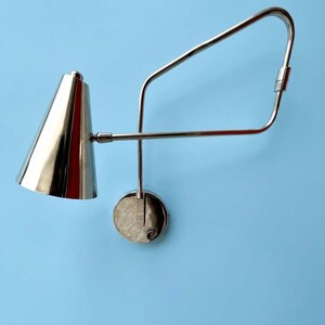 May include: A contemporary wall lamp in a silver finish, featuring a conical lampshade and an adjustable arm. The lamp is mounted on a circular base. The background is a light blue colour.