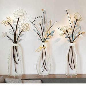 May include: A set of three metal wall art pieces, each featuring a stylised vase containing a bouquet of flowers. The designs incorporate wire-like stems and leaves in shades of gold, light blue, and white, creating a delicate floral arrangement.