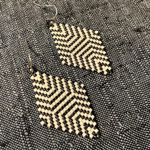 May include: A pair of handmade earrings with a geometric pattern. The earrings are diamond-shaped, featuring a black and cream-colored zigzag design. The earrings have silver-colored ear wires and are displayed on a textured gray surface.