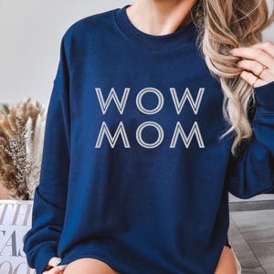 Könnte beinhalten: Ein marineblaues Sweatshirt mit dem weißen Schriftzug "WOW MOM" in umrandeten Buchstaben. Das Bild zeigt auch eine dekorative Anordnung aus getrockneten Pflanzen und ein Buch.