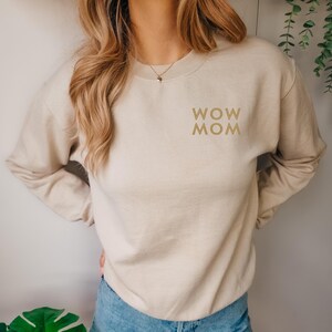 Könnte beinhalten: Beiges Sweatshirt mit dem goldenen Schriftzug „WOW MOM“. Das Oberteil mit Rundhalsausschnitt und langen Ärmeln wird mit einer blauen Jeans kombiniert. Das Sweatshirt im lässigen Stil ist aus einem weichen Material gefertigt.