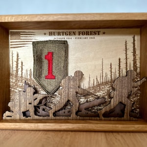Puede incluir: Una caja de sombra de madera con una escena de bosque grabada con láser y tres siluetas de soldados. Un parche verde oliva con un número uno rojo está centrado. El texto dice HURTGEN FOREST OCTOBER 1944 - FEBRUARY 1945 con una bandera de EE. UU.