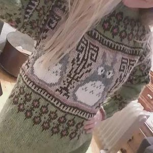 May include: A green knitted sweater with a detailed pattern. The sweater features a central design with two cartoon characters, surrounded by geometric and floral patterns in shades of brown, pink, and white. The sleeves and lower portion of the sweater are a solid green.