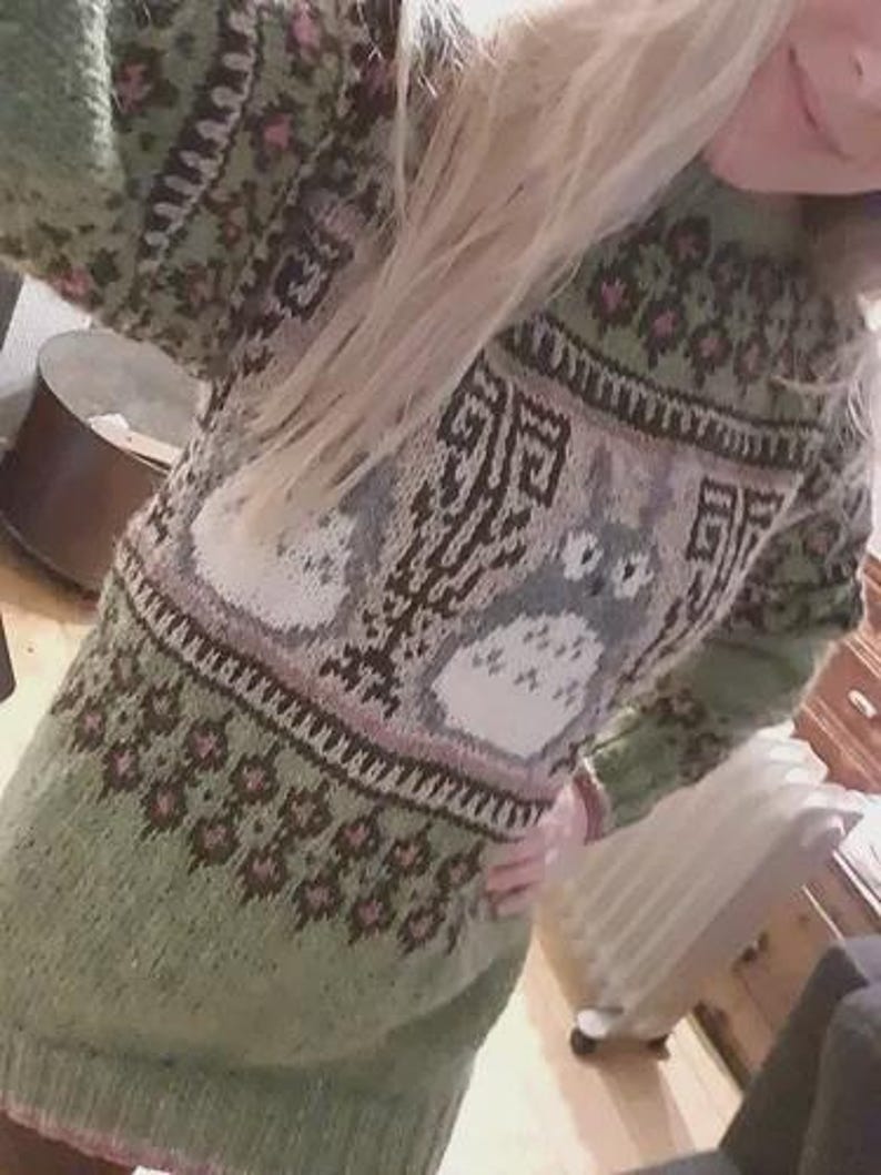 May include: A green knitted sweater with a detailed pattern. The sweater features a central design with two cartoon characters, surrounded by geometric and floral patterns in shades of brown, pink, and white. The sleeves and lower portion of the sweater are a solid green.