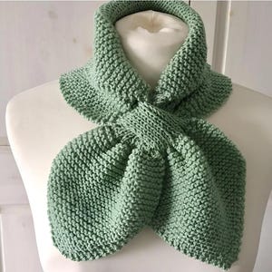 Puede incluir: Bufanda de punto verde salvia exhibida en un maniquí blanco. La bufanda está hecha de un hilo grueso y texturizado y está anudada en la parte delantera. La bufanda es de color liso y parece hecha a mano. Es un accesorio de invierno.