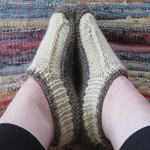 Puede incluir: Calcetines de pantuflas tejidos a mano en crema y marrón. Los calcetines presentan un patrón de puntadas texturizadas y cubren los pies y los tobillos. Los calcetines se muestran en los pies de una persona, descansando sobre una alfombra multicolor.