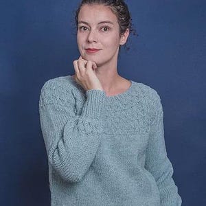 Puede incluir: Un suéter azul claro con cuello redondo y mangas largas. El suéter tiene un patrón texturizado en la parte superior del pecho y las mangas. La persona de la imagen lleva el suéter y tiene la mano en la barbilla.