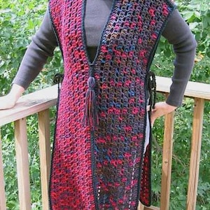 May include: A long, open-front crochet vest in shades of red and black. The vest features a deep V-neck, a tassel detail, and side ties. The open weave design is visible, and the vest appears to be handmade.