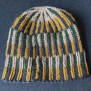 Könnte beinhalten: Eine gestrickte Beanie-Mütze mit vertikalen Streifen in den Farben Gelb, Grün, Blau und Creme. Die Mütze ist aus einem weichen, strukturierten Garn gefertigt und hat einen gerippten Bund.