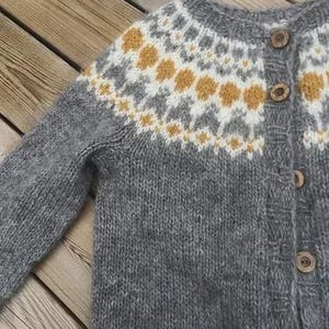 May include: A gray knit cardigan with a patterned yoke featuring white, gray, and yellow accents. The sweater has wooden buttons and long sleeves. The garment is made of a soft, textured yarn.