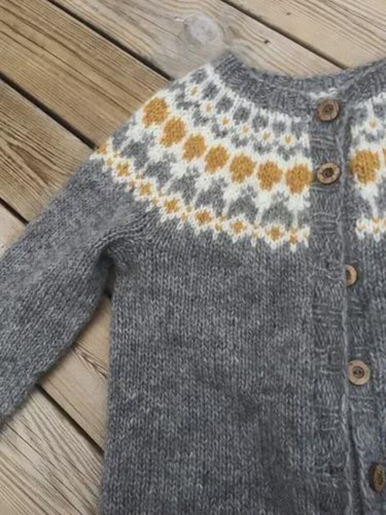 May include: A gray knit cardigan with a patterned yoke featuring white, gray, and yellow accents. The sweater has wooden buttons and long sleeves. The garment is made of a soft, textured yarn.