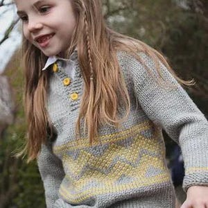 Pode incluir: Um suéter cinza de malha com um padrão de chevron amarelo. O suéter tem uma gola abotoada e é usado com jeans azuis. É uma peça de roupa infantil.