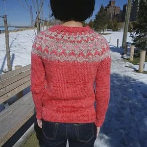 May include: A coral-colored knit sweater with a patterned yoke in white and pink. The sweater has long sleeves and a crew neck. The person is wearing dark blue jeans. The background shows a snowy landscape and a clear blue sky.