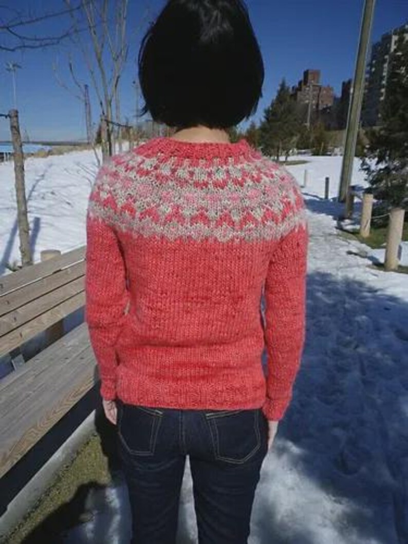 May include: A coral-colored knit sweater with a patterned yoke in white and pink. The sweater has long sleeves and a crew neck. The person is wearing dark blue jeans. The background shows a snowy landscape and a clear blue sky.