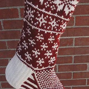 Puede incluir: Un calcetín de Navidad tejido en burdeos y blanco, con un diseño estampado. La puntera y el talón son blancos, y el puño es acanalado. El calcetín cuelga contra una pared de ladrillos rojos.