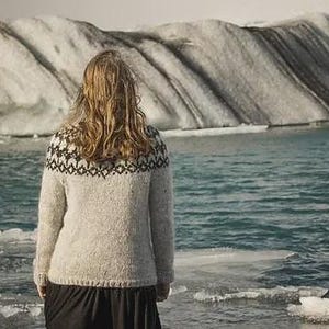 May include: A person wearing a light gray sweater with a black and white patterned yoke, standing near a body of water with ice floes. The background features a glacier and a body of water.