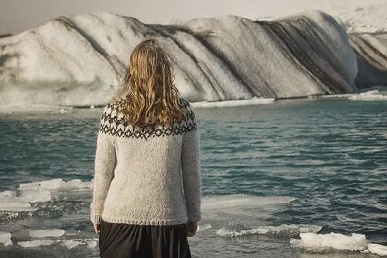 May include: A person wearing a light gray sweater with a black and white patterned yoke, standing near a body of water with ice floes. The background features a glacier and a body of water.