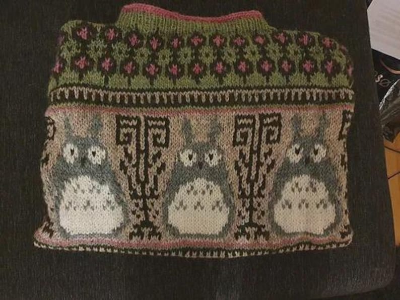 May include: A knitted sweater featuring a design of three cartoon characters with large eyes and white bellies. The sweater has a green and pink floral pattern at the top, with a pink neckline and cuffs. The main colors are gray, white, black, green, and pink.