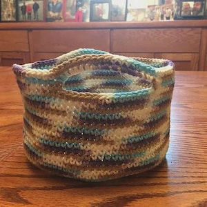 May include: A handmade crochet basket in shades of blue, purple, brown, and cream. The basket has two handles and is sitting on a wooden table. The yarn has a slight shimmer, adding a touch of elegance to this storage solution.