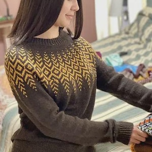 May include: A dark brown sweater with a geometric yellow pattern across the chest and shoulders. The sweater has a crew neck and long sleeves. The person is holding a stack of folded textiles.