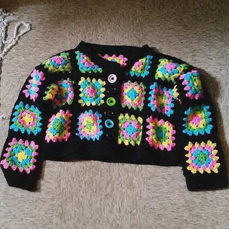 May include: Black crochet cardigan with colorful square patterns. The squares feature pink, yellow, blue, and green accents. The cardigan has a round neckline and is buttoned with various colored buttons.