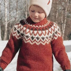 Può includere: Un bambino indossa un maglione rosso lavorato a maglia con un motivo geometrico bianco e marrone intorno al giromanica. Il bambino indossa anche un cappello di lana color crema con un pompon e un piccolo accento rosso. Lo sfondo è una scena esterna innevata.