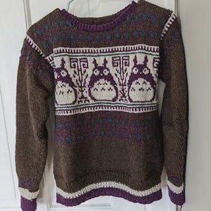May include: Brown knit sweater with a purple and white pattern. The sweater features a repeating design of cartoon characters across the chest. The cuffs and hem are trimmed with purple and white stripes. The sweater is hanging on a white door.