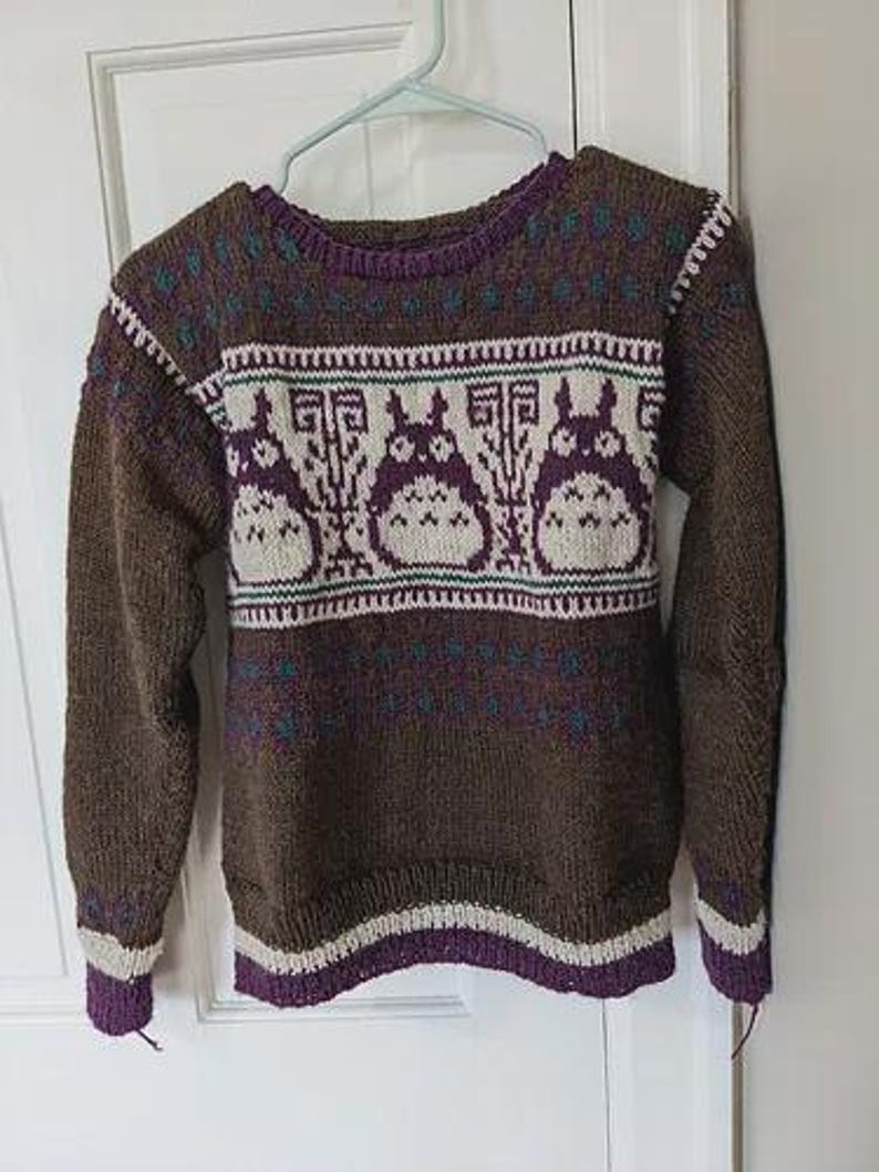 May include: Brown knit sweater with a purple and white pattern. The sweater features a repeating design of cartoon characters across the chest. The cuffs and hem are trimmed with purple and white stripes. The sweater is hanging on a white door.