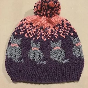 Può includere: Un berretto lavorato a maglia nei toni del viola, grigio e rosa. Il cappello presenta un motivo di gatti grigi con colletti rosa su uno sfondo viola. La parte superiore del cappello è rosa e viola scuro con un pompon abbinato.