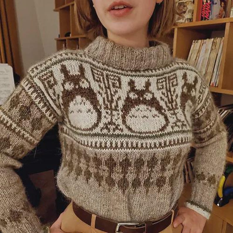 May include: A beige and brown knit sweater with a high collar. The sweater features a repeating pattern of cartoon characters. The sleeves and lower portion of the sweater have a geometric design. The sweater is worn with a brown belt.