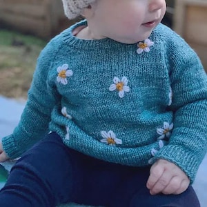 Può includere: Un maglione lavorato a maglia color verde acqua ornato da applicazioni di margherite bianche e gialle. Il maglione ha maniche lunghe e viene indossato con pantaloni blu navy. Un capo comodo ed elegante per bambini.