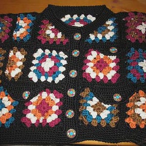 May include: A black crochet cardigan featuring colorful square patterns in shades of white, orange, blue, and burgundy. The cardigan has a round neckline and is fastened with decorative buttons. The garment is handmade and has a vintage aesthetic.