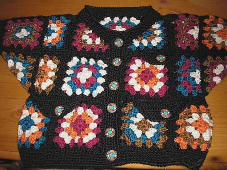 May include: A black crochet cardigan featuring colorful square patterns in shades of white, orange, blue, and burgundy. The cardigan has a round neckline and is fastened with decorative buttons. The garment is handmade and has a vintage aesthetic.
