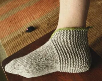 May include: A gray knitted sock with a green trim around the ankle. The sock is on a person's foot, resting on a wooden surface. A small black toy mouse with a pink nose is visible in the background.